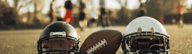 Two football helmets and a football resting on a grassy field with blurred players practicing in the background during a team workout.