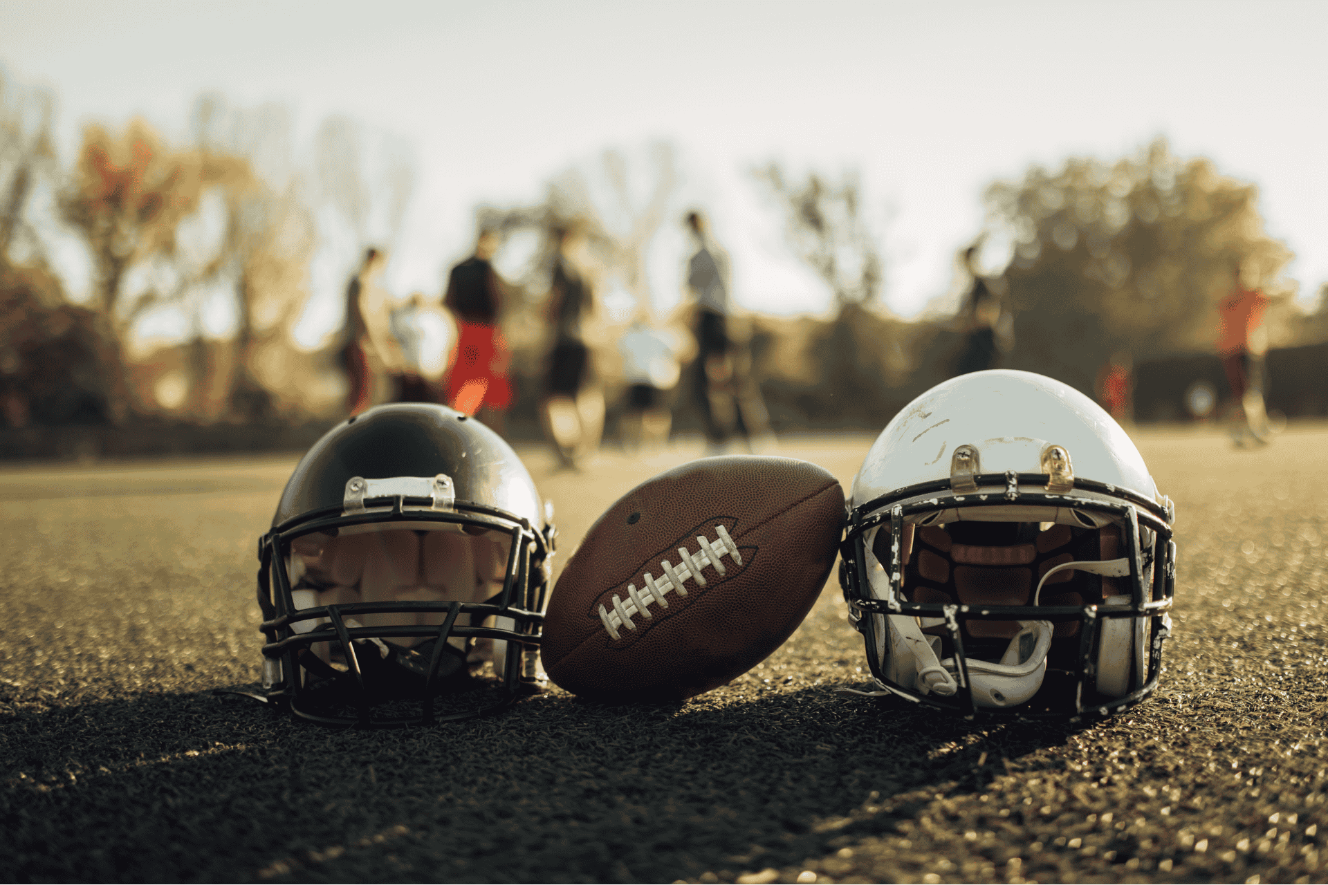 Two football helmets and a football resting on a grassy field with blurred players practicing in the background during a team workout.