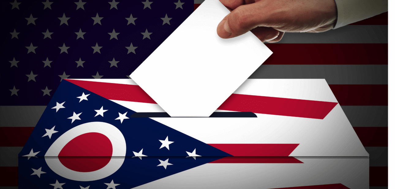 A close-up of a hand placing a white ballot paper into a ballot box, featuring a graphic of the Ohio state flag and the United States flag.