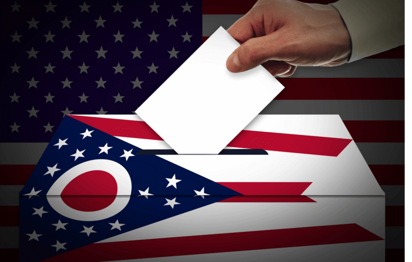 A close-up of a hand placing a white ballot paper into a ballot box, featuring a graphic of the Ohio state flag and the United States flag.