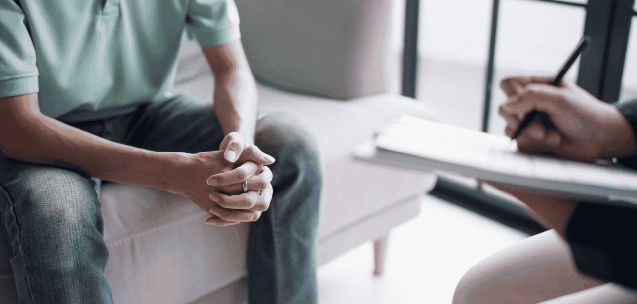 A patient sits with hands clasped during a therapy session with a mental health professional taking notes on a clipboard.
