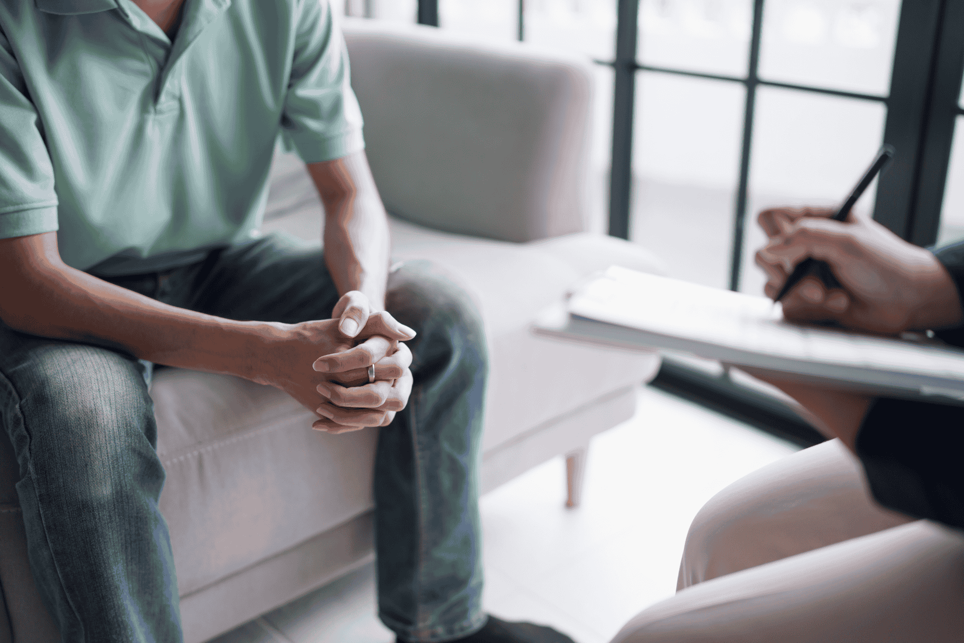 A patient sits with hands clasped during a therapy session with a mental health professional taking notes on a clipboard.