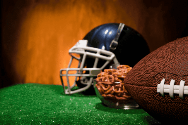 A football helmet and a leather football rest on green turf against a blurred orange background.