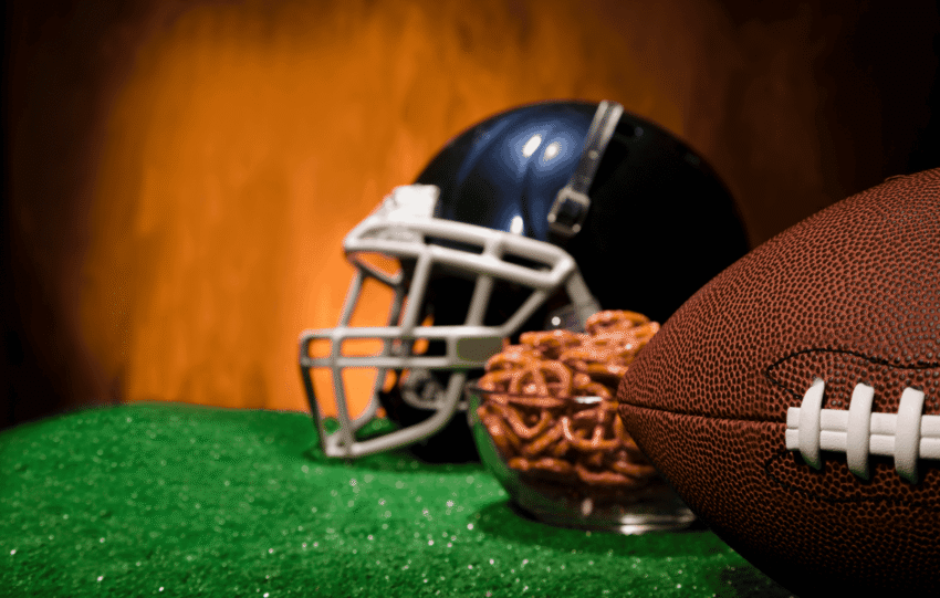 A football helmet and a leather football rest on green turf against a blurred orange background.
