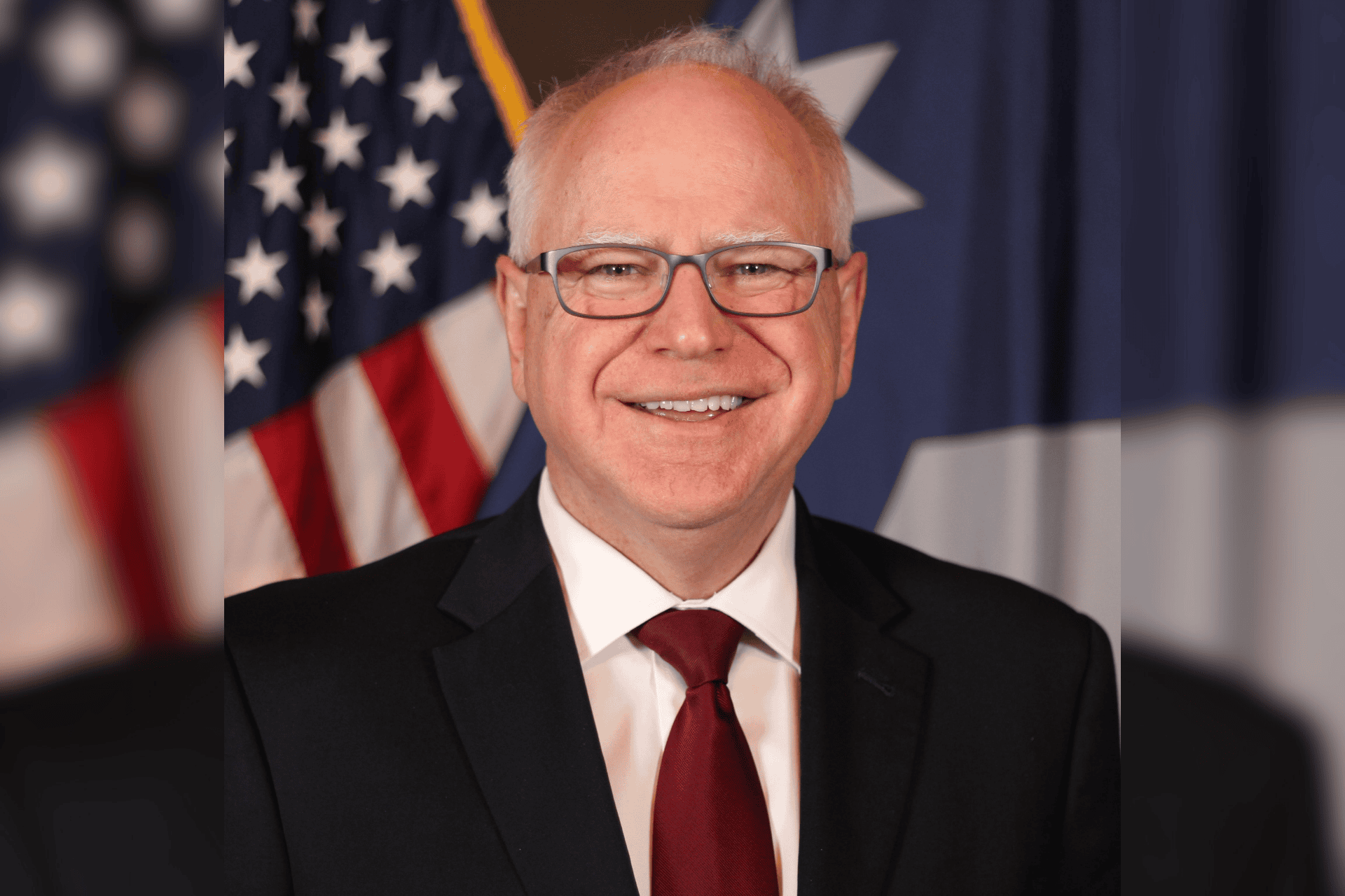 Minnesota Governor Tim Walz smiling in a suit in front of American flags.