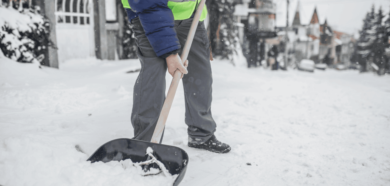 A person wearing a winter coat and high-visibility vest shoveling deep snow off a sidewalk during a winter storm in the Cincinnati Tri-State area.