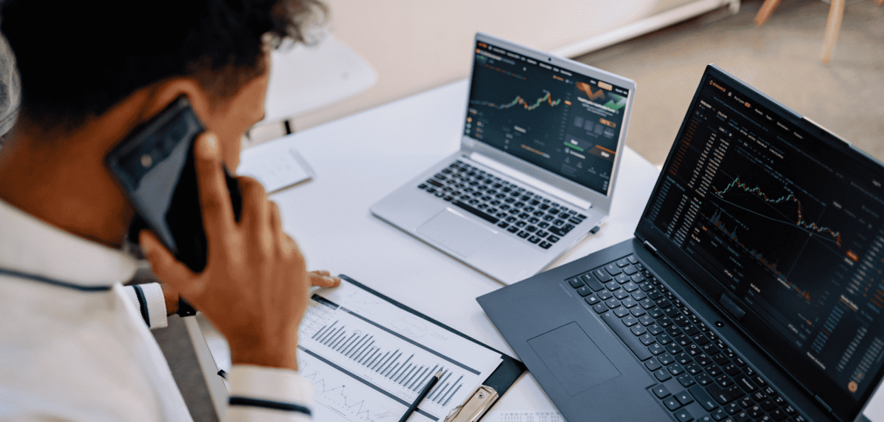 A person in a white shirt sits at a desk in a bright office, talking on a smartphone while looking at multiple laptop screens displaying data charts and graphs.
