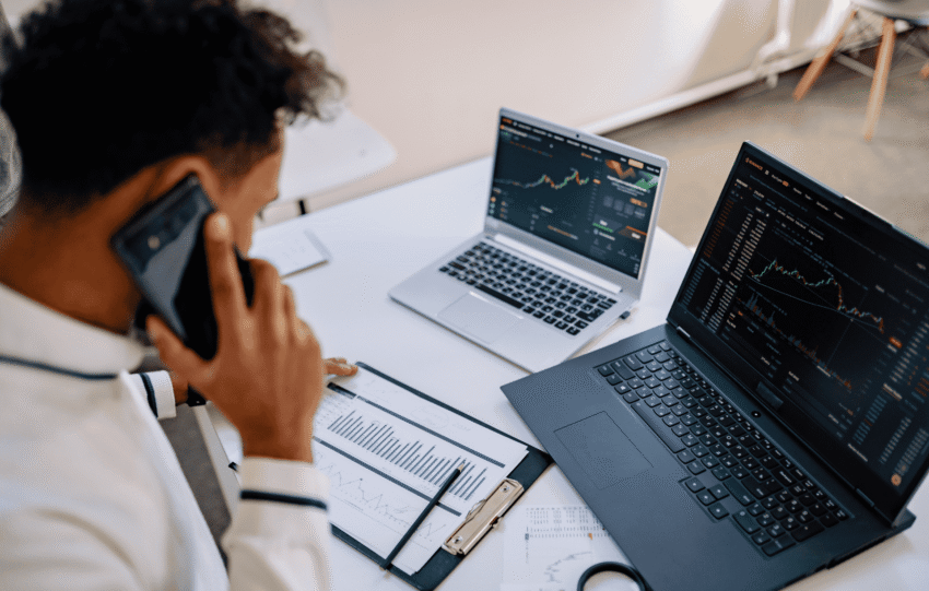 A person in a white shirt sits at a desk in a bright office, talking on a smartphone while looking at multiple laptop screens displaying data charts and graphs.