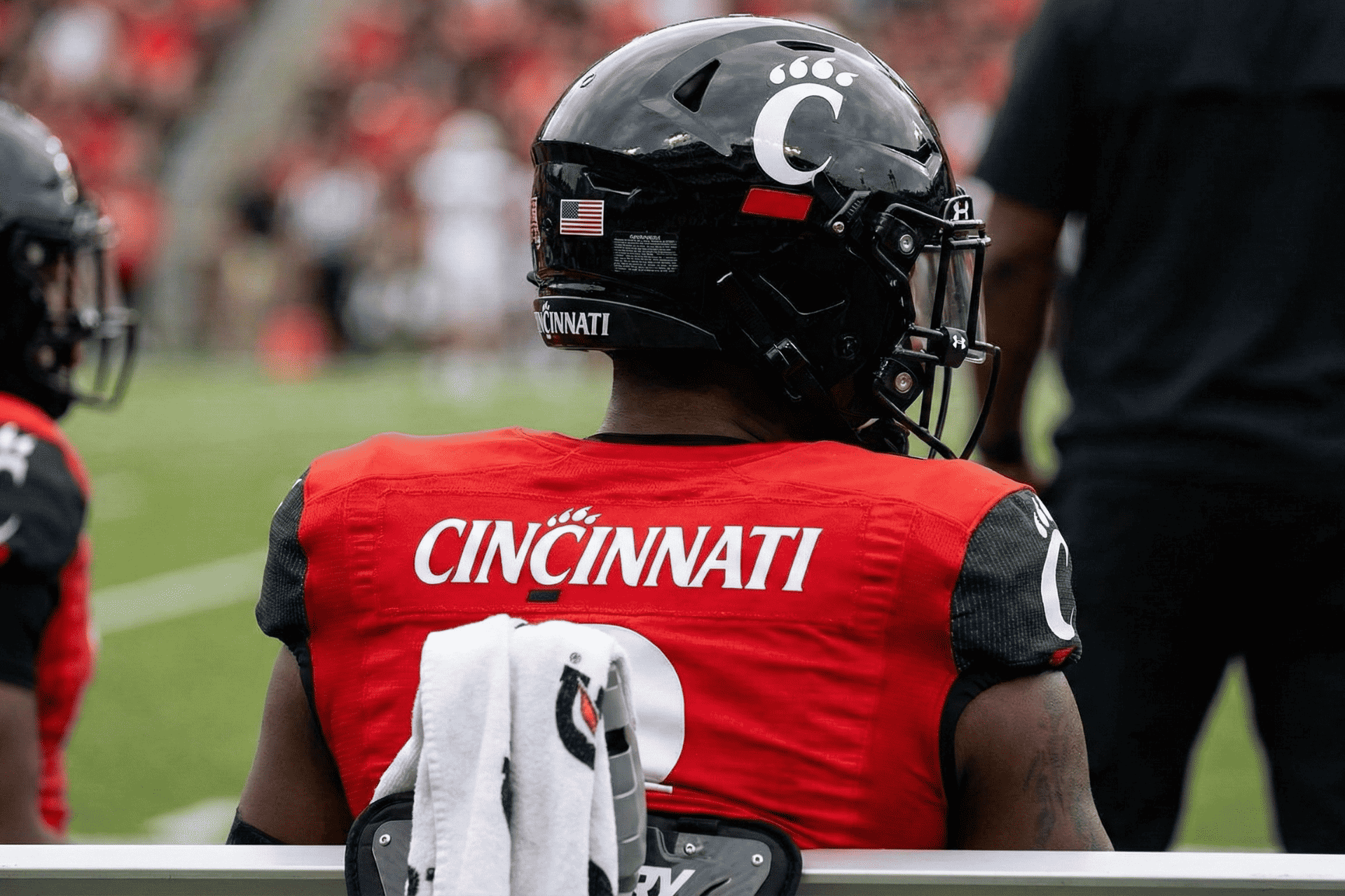 Rear view of a Cincinnati Bearcats football player wearing a red jersey and black helmet looking out onto the field.