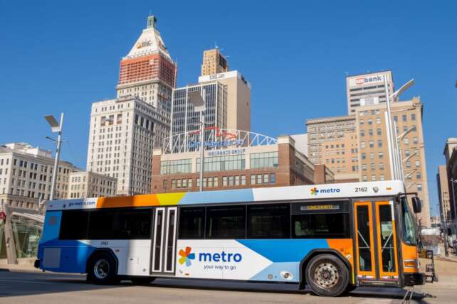 A blue and white Cincinnati Metro bus driving through downtown Cincinnati with the city skyline in the background on a sunny day.