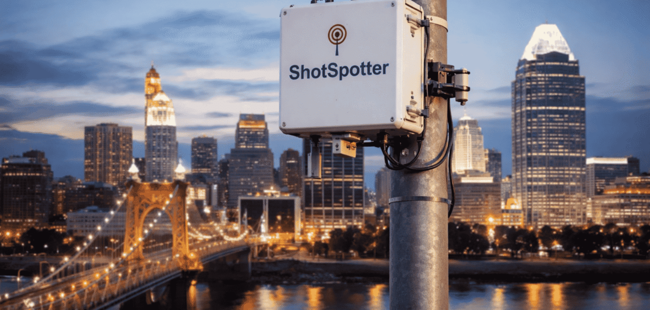 ShotSpotter gunshot detection sensor mounted on a pole with the Cincinnati skyline and Roebling Bridge in the background.