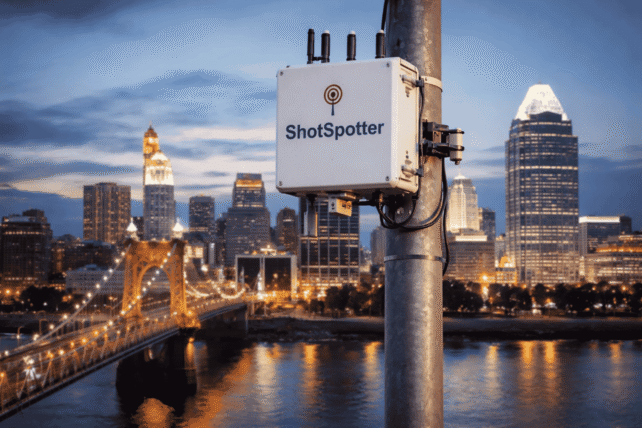 ShotSpotter gunshot detection sensor mounted on a pole with the Cincinnati skyline and Roebling Bridge in the background.