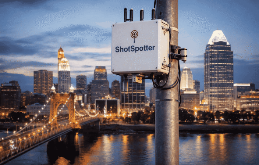 ShotSpotter gunshot detection sensor mounted on a pole with the Cincinnati skyline and Roebling Bridge in the background.