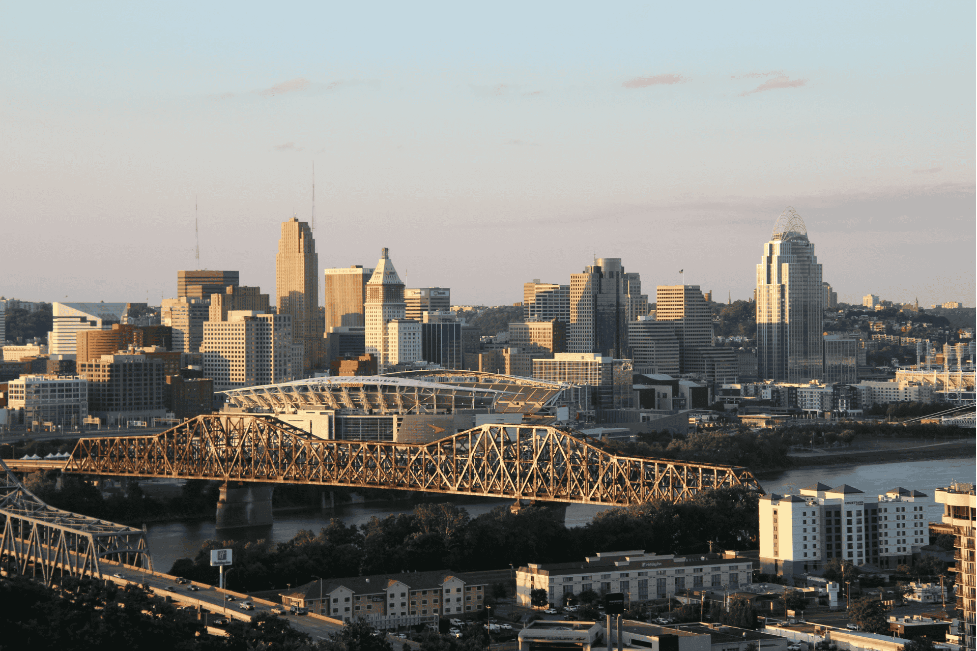 Aerial view of the Cincinnati, Ohio skyline featuring the John A. Roebling Suspension Bridge, the Ohio River, and downtown skyscrapers during the day.