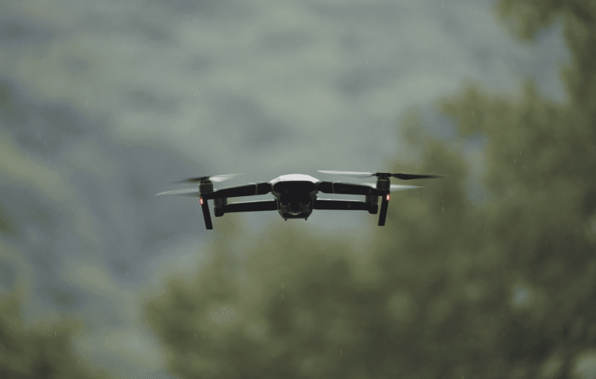 A small black quadcopter drone hovering in mid-air against a blurred background of trees and sky, illustrating defense drone production in Ohio.