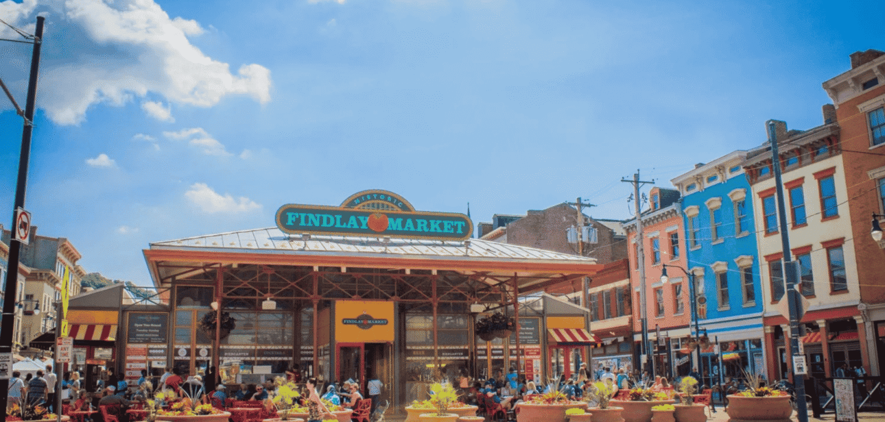 Busy outdoor dining scene at Findlay Market in Cincinnati with colorful historic buildings. Text overlay reads: "Findlay Market Cincinnati fuels tourism with vibrant local food culture."