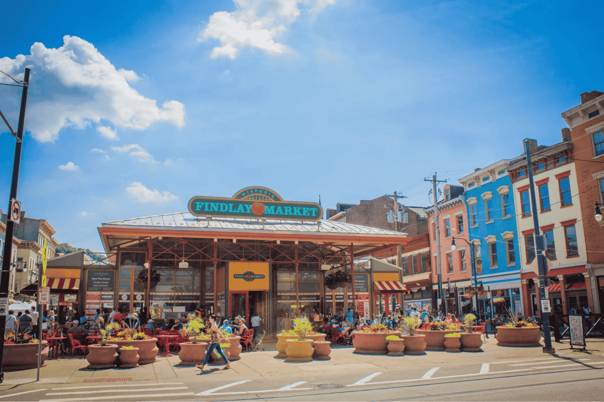 Busy outdoor dining scene at Findlay Market in Cincinnati with colorful historic buildings. Text overlay reads: "Findlay Market Cincinnati fuels tourism with vibrant local food culture."
