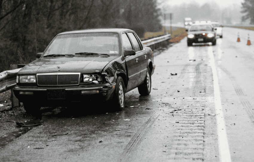 A dark, heavily damaged sedan stopped on a wet highway with a police vehicle in the background, related to the hit-and-run investigation involving a Cincinnati congressional candidate.
