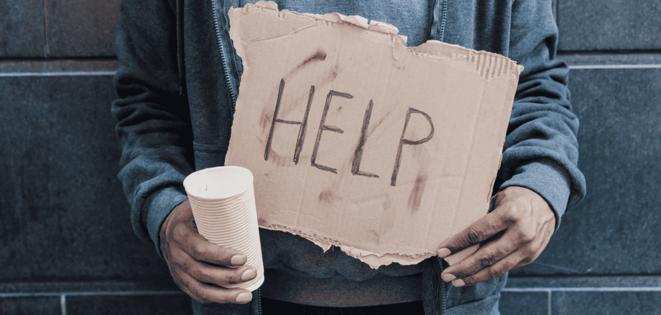 A person wearing a gray jacket stands against a dark wall, holding a white cup and a cardboard sign that reads "HELP".
