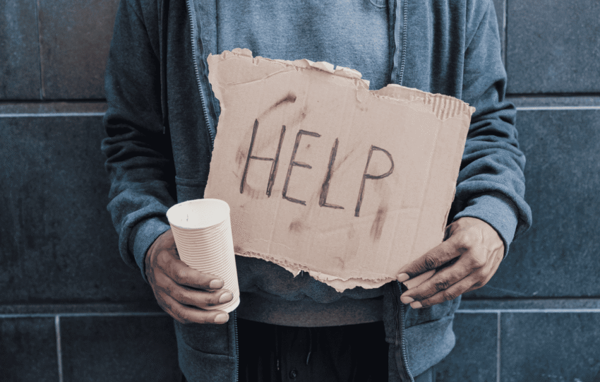 A person wearing a gray jacket stands against a dark wall, holding a white cup and a cardboard sign that reads "HELP".