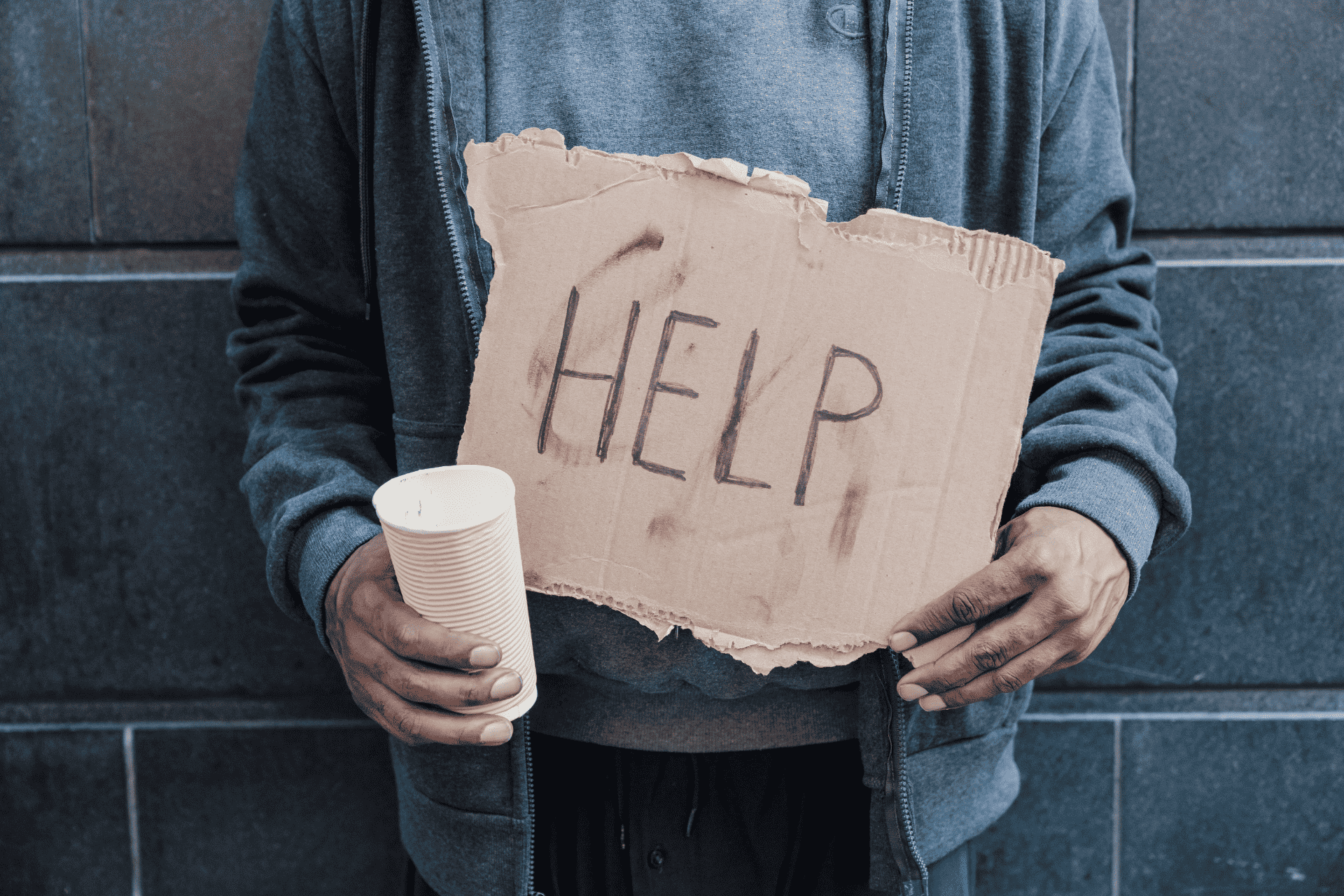 A person wearing a gray jacket stands against a dark wall, holding a white cup and a cardboard sign that reads "HELP".