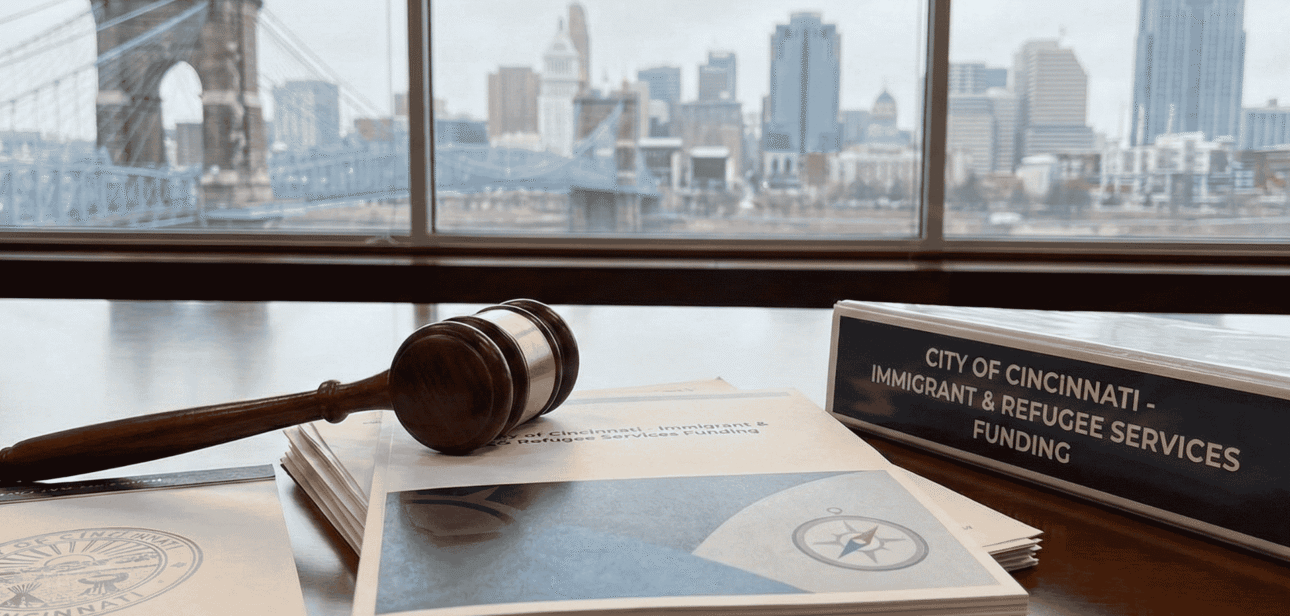 A wooden gavel and official documents resting on a desk with the Cincinnati skyline and Roebling Suspension Bridge visible through a window in the background.