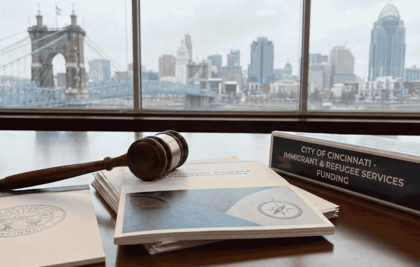 A wooden gavel and official documents resting on a desk with the Cincinnati skyline and Roebling Suspension Bridge visible through a window in the background.