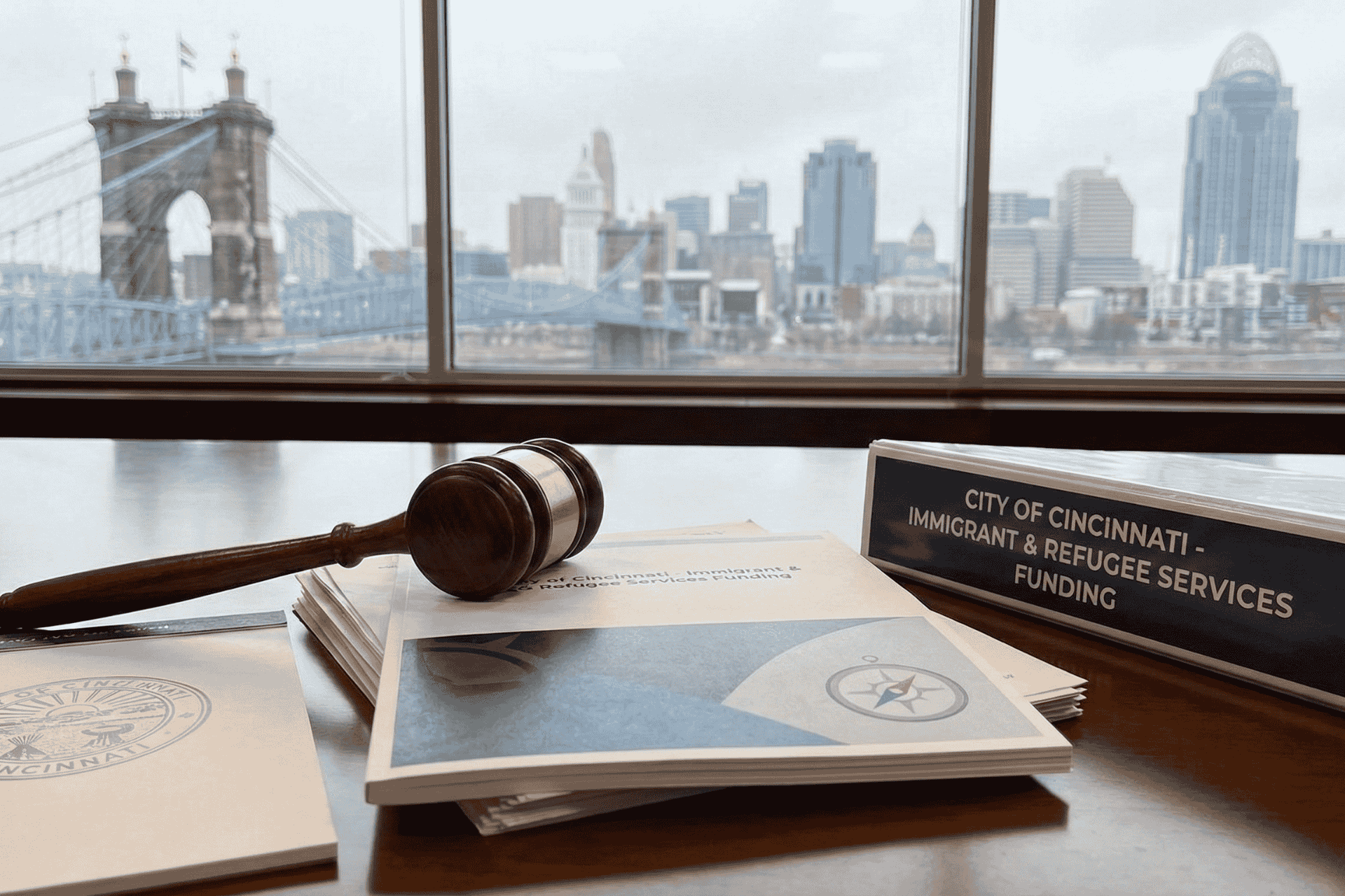 A wooden gavel and official documents resting on a desk with the Cincinnati skyline and Roebling Suspension Bridge visible through a window in the background.