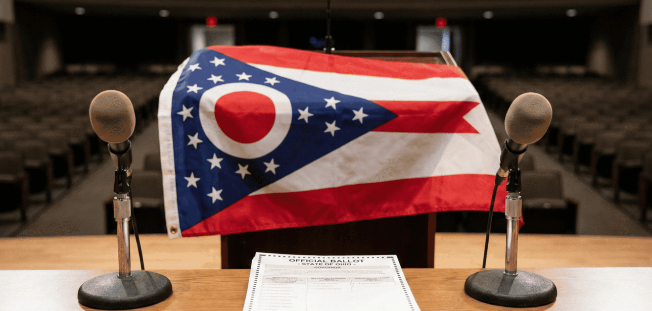 A wooden podium with microphones stands in front of the Ohio state flag, representing the open seat race for the 2026 Ohio Governor's election.