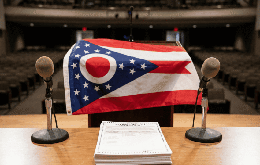 A wooden podium with microphones stands in front of the Ohio state flag, representing the open seat race for the 2026 Ohio Governor's election.