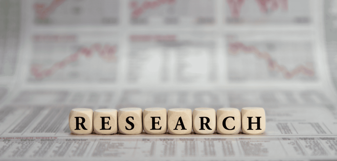 Wooden letter blocks spelling "RESEARCH" sitting on top of blurred newspapers and statistical data sheets.