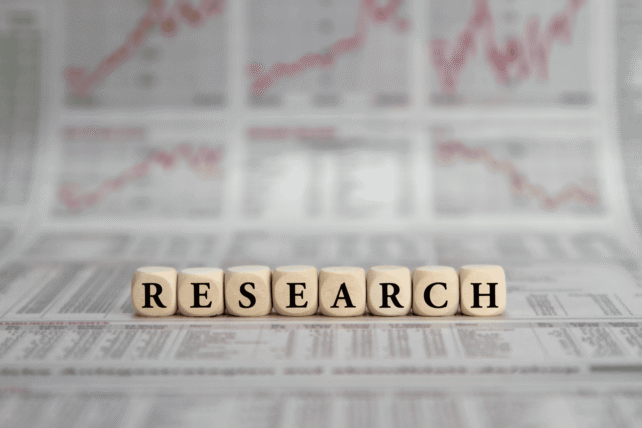 Wooden letter blocks spelling "RESEARCH" sitting on top of blurred newspapers and statistical data sheets.