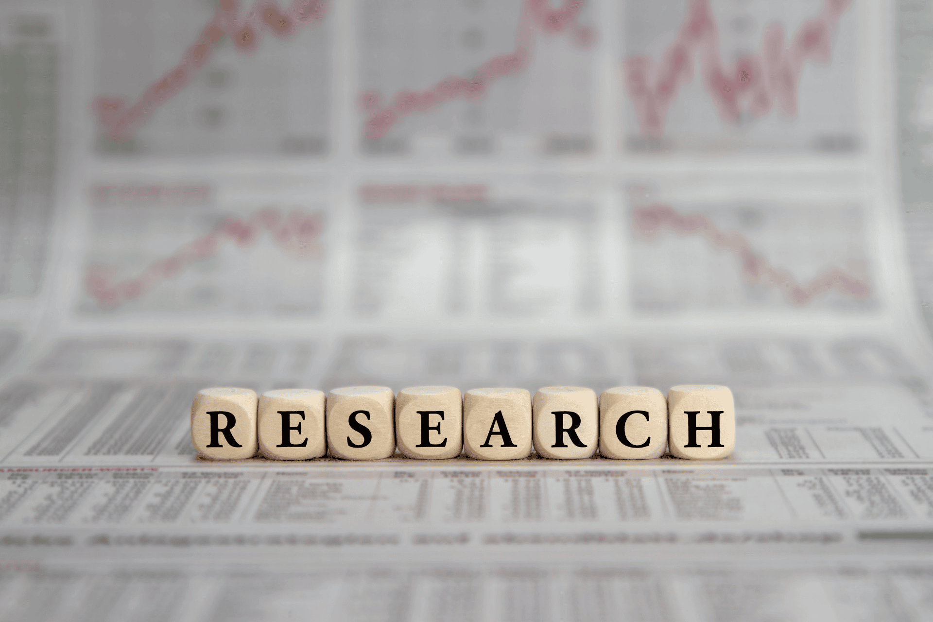 Wooden letter blocks spelling "RESEARCH" sitting on top of blurred newspapers and statistical data sheets.