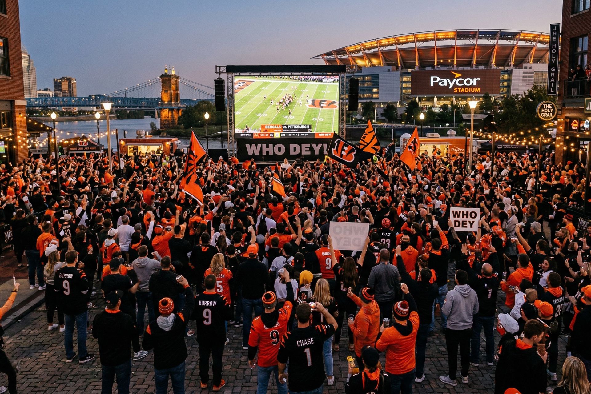 A large crowd of Cincinnati Bengals fans in orange and black apparel gathers at The Banks to watch a game on a massive outdoor LED screen, with Paycor Stadium and the Roebling Bridge visible under a twilight sky.