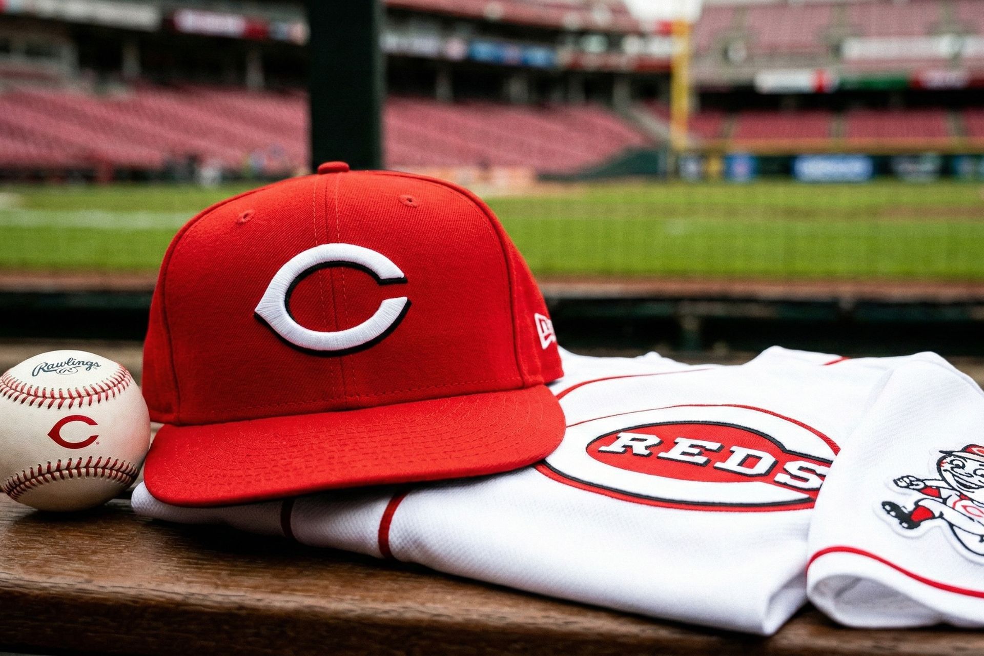 A red Cincinnati Reds baseball cap resting on a folded white home jersey on a ledge overlooking the field at Great American Ball Park.