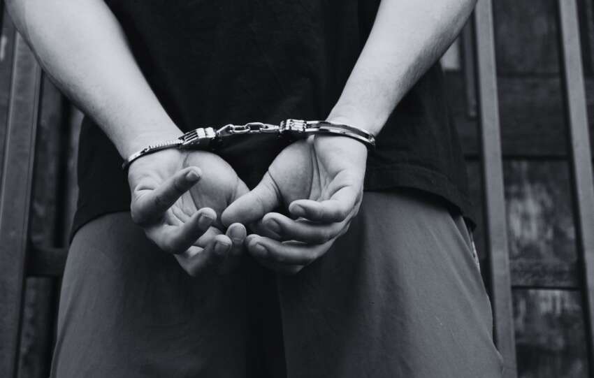 A close-up, black-and-white image of a person's hands secured in handcuffs behind their back.