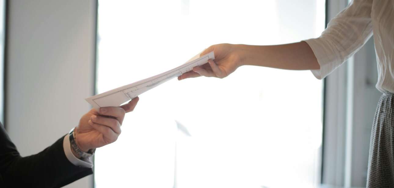 A job applicant hands a printed resume to an employer across a desk.
