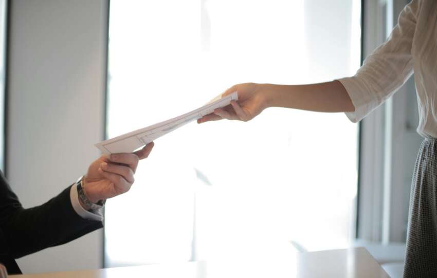 A job applicant hands a printed resume to an employer across a desk.