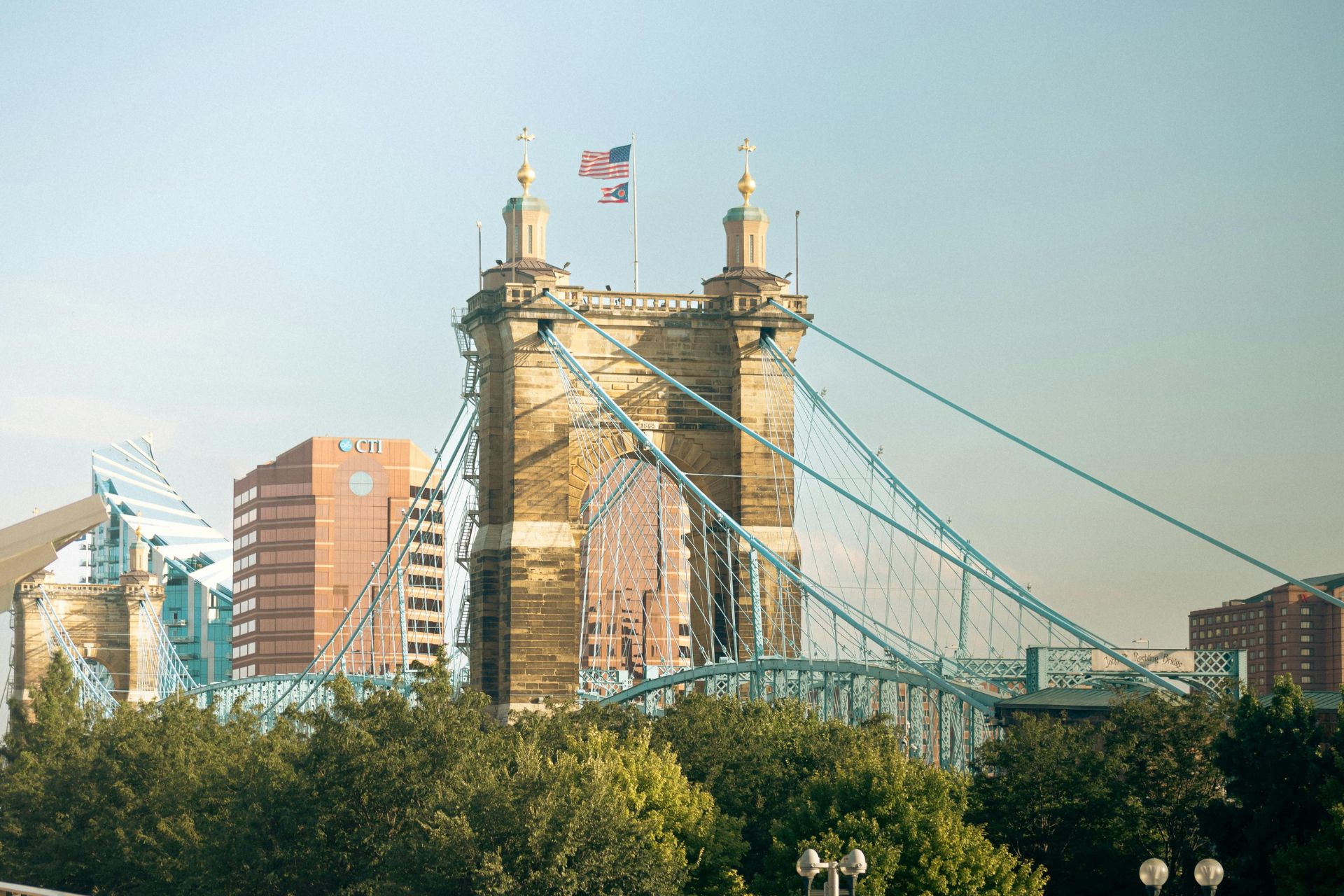 Cincinnati, Ohio skyline featuring the historic blue John A. Roebling Suspension Bridge spanning the Ohio River under a blue sky.