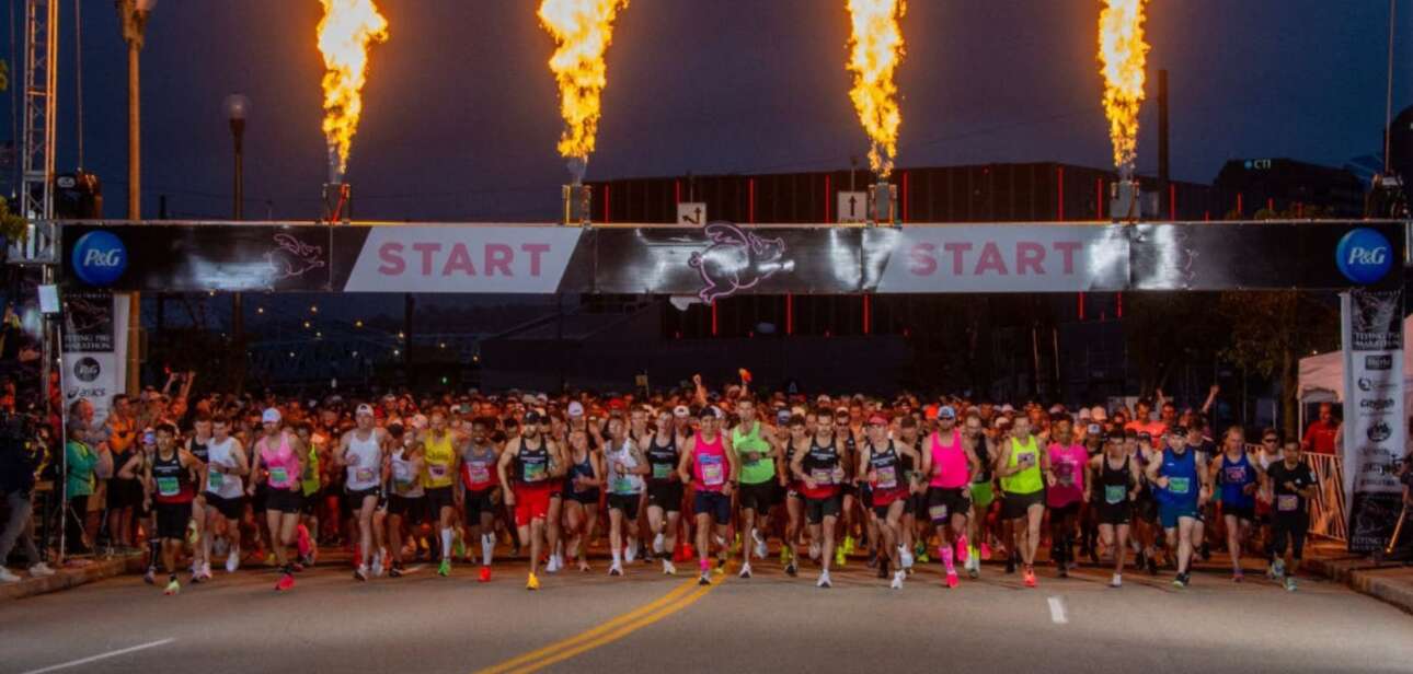 Runners gathered behind the start line banner of the Cincinnati Flying Pig Marathon in the early morning, with large ceremonial flames shooting from towers on either side of the street.