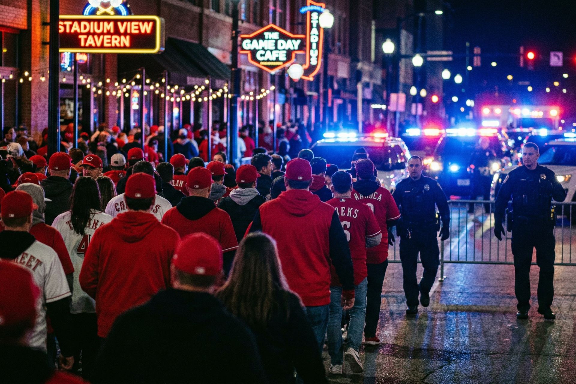 Tense nighttime scene at The Banks in Cincinnati after Opening Day 2026, showing a large crowd of fans in red apparel facing a line of police officers and flashing emergency lights.