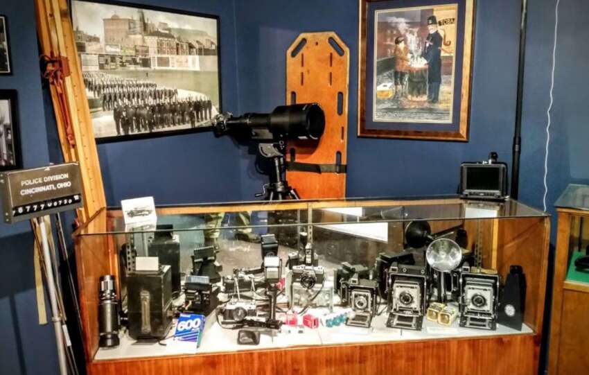 A display case at the Greater Cincinnati Police Museum featuring historical law enforcement artifacts, including vintage cameras, radios, and a tabletop siren.