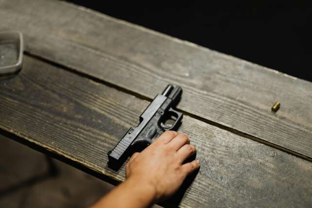 A person's hand resting near a black handgun on a dark wooden surface.