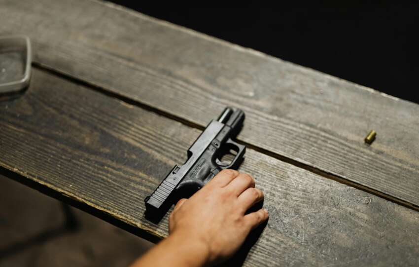 A person's hand resting near a black handgun on a dark wooden surface.