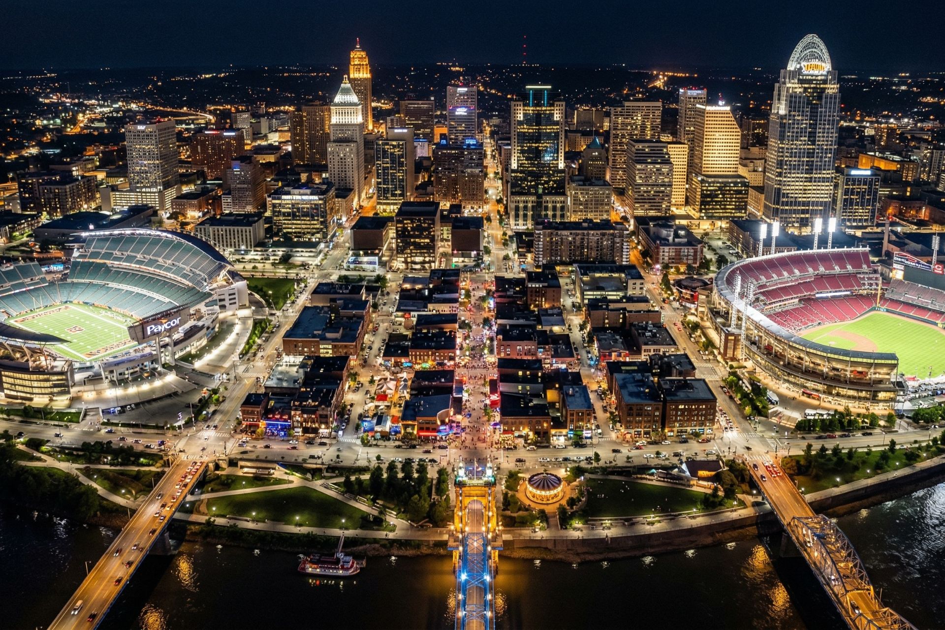 Aerial bird's-eye view of The Banks entertainment district in Cincinnati at night, showing the illuminated Paycor Stadium and Great American Ball Park along the Ohio River.