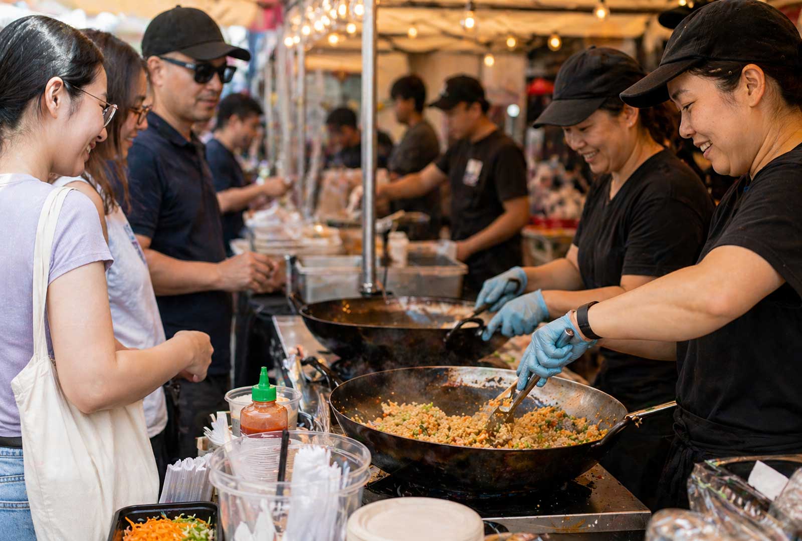 Asian food vendors cooking fried rice in large woks while a line of customers waits at a busy festival booth
