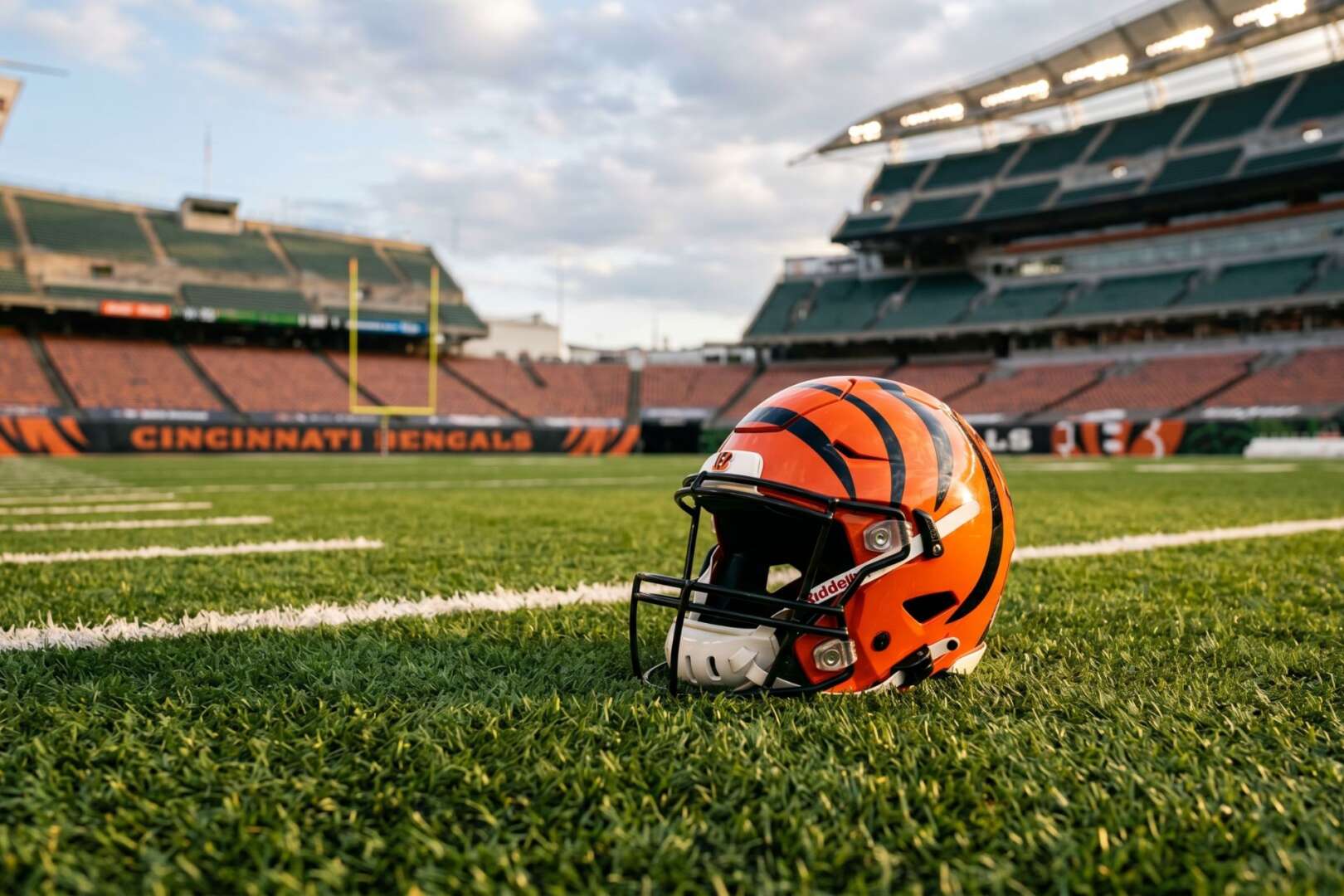 A Cincinnati Bengals football helmet sits on the grass of Paycor Stadium field, with the empty orange and black stands and stadium lights in the background.