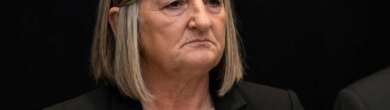 Close-up of Teresa Theetge with a tense expression, wearing a black blazer and white blouse against a dark background following her firing as Cincinnati police chief