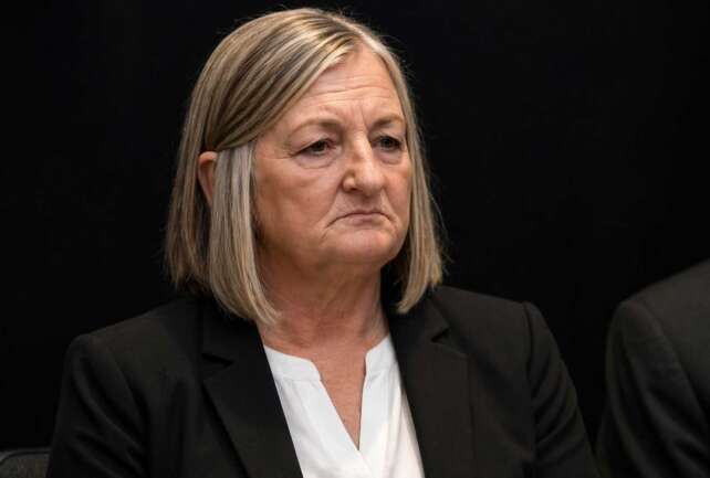 Close-up of Teresa Theetge with a tense expression, wearing a black blazer and white blouse against a dark background following her firing as Cincinnati police chief