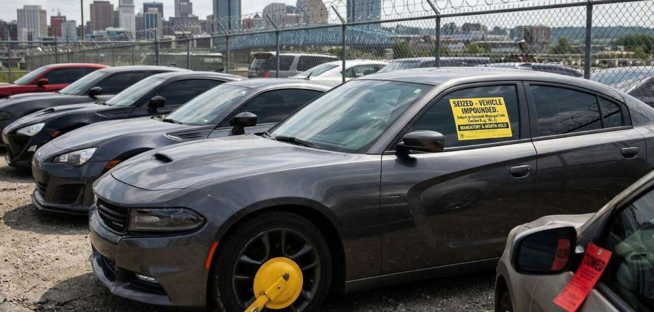 Row of impounded vehicles with a yellow wheel boot and seized window sticker in a fenced dirt lot, with the Cincinnati skyline in the background, representing proposed penalties for Cincinnati street takeovers.